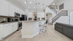 A modern kitchen with white cabinetry, a central island, bar stools, and a staircase leading to an upper level. - Theater Room | Games Area | Kids Themed Bedrooms | Private Pool (Kissimmee)