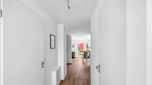 Un couloir aux murs blancs et au plancher en bois mène à une pièce dont un mur est peint en rouge et qui possède une porte.
