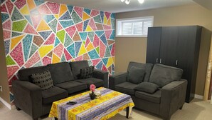 Ein Wohnzimmer mit zwei grauen Sofas, einer Wand mit farbigem geometrischen Muster, einem kleinen Tisch mit floralem Tischtuch und einem dunklen Holzschrank.