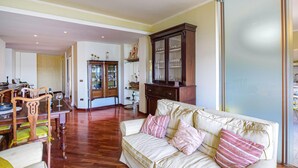 A spacious living room with a wooden dining table, chairs, a cream-colored sofa with striped cushions, and a dark wooden cabinet with glass doors. - 2 bedroom cozy apartment in Orbetello (Orbetello)