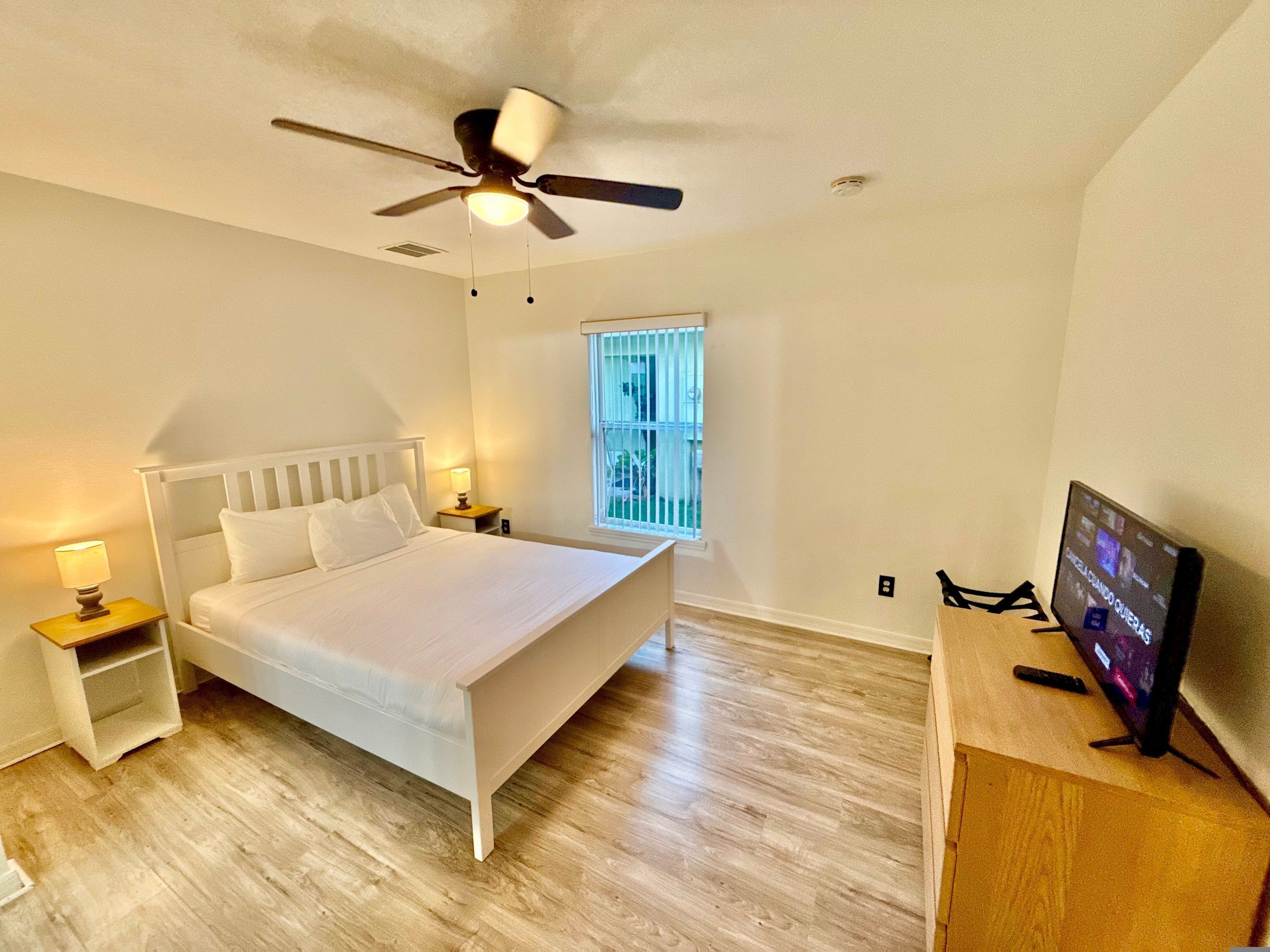 A bedroom with a bed, nightstands, a ceiling fan, a TV, and a window with blinds.