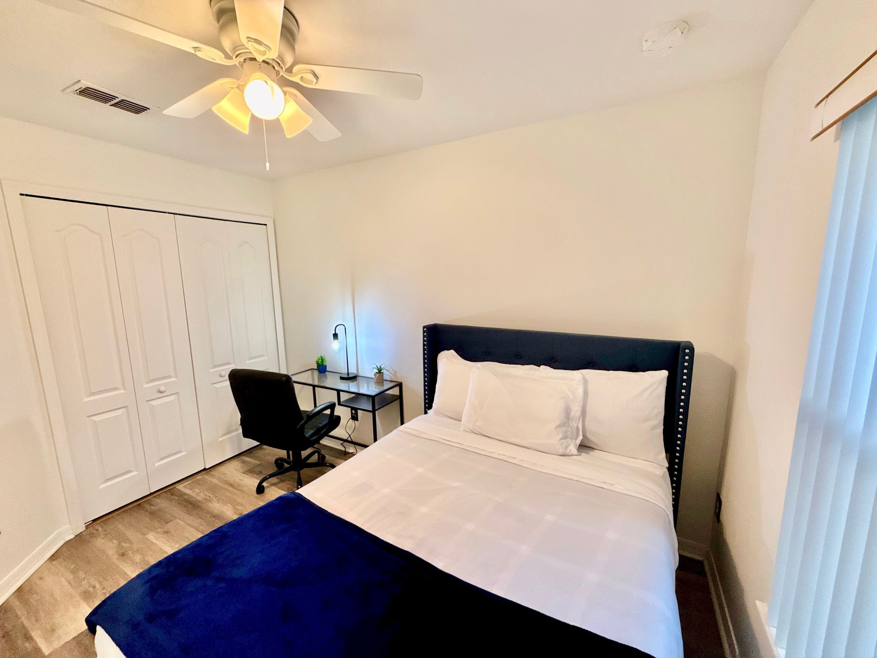 A bedroom with a bed, a desk, a chair, a ceiling fan, and a door.