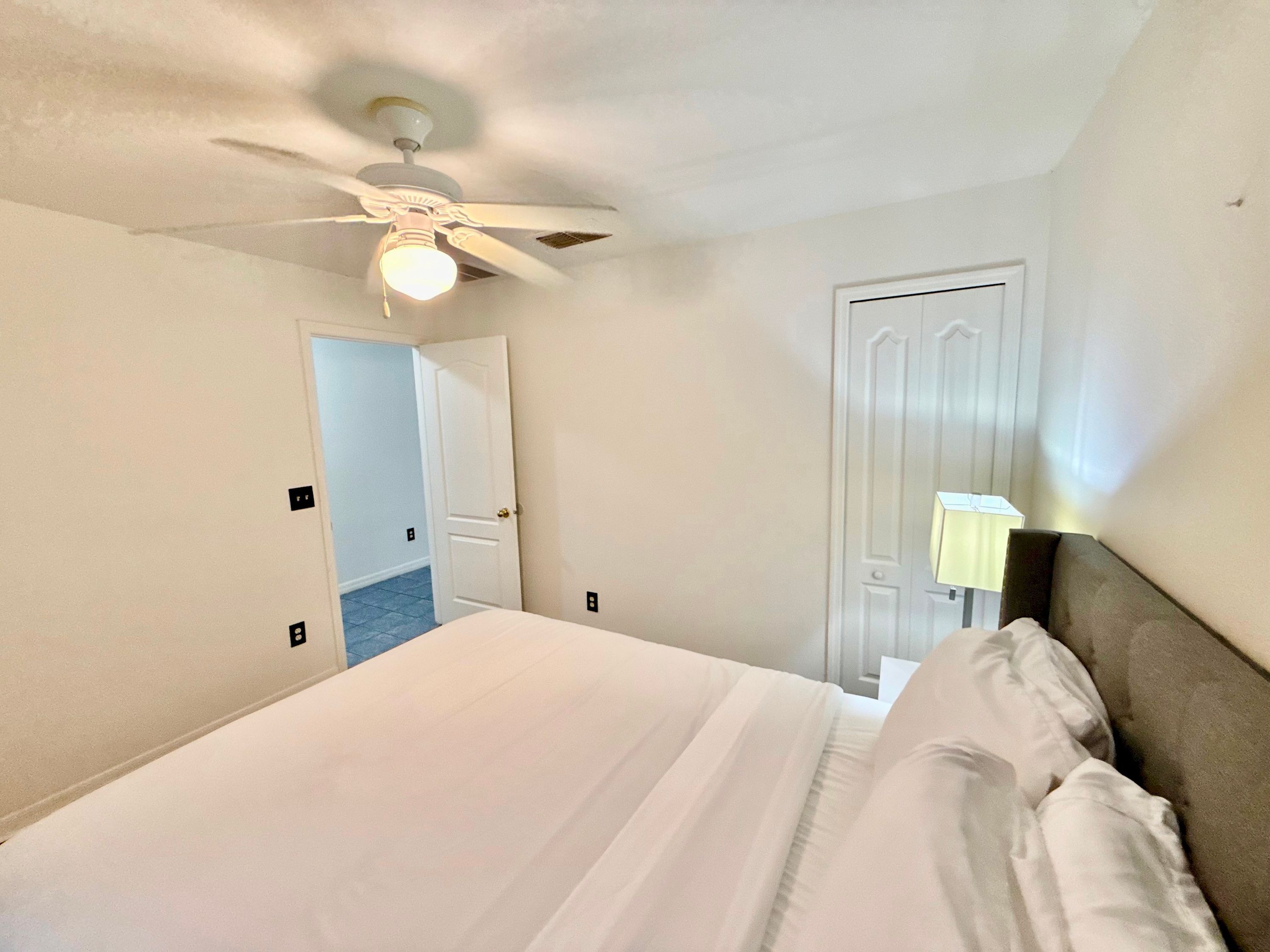 Une chambre d’hôtel avec un lit, un ventilateur de plafond, une porte et une table de chevet avec une lampe.