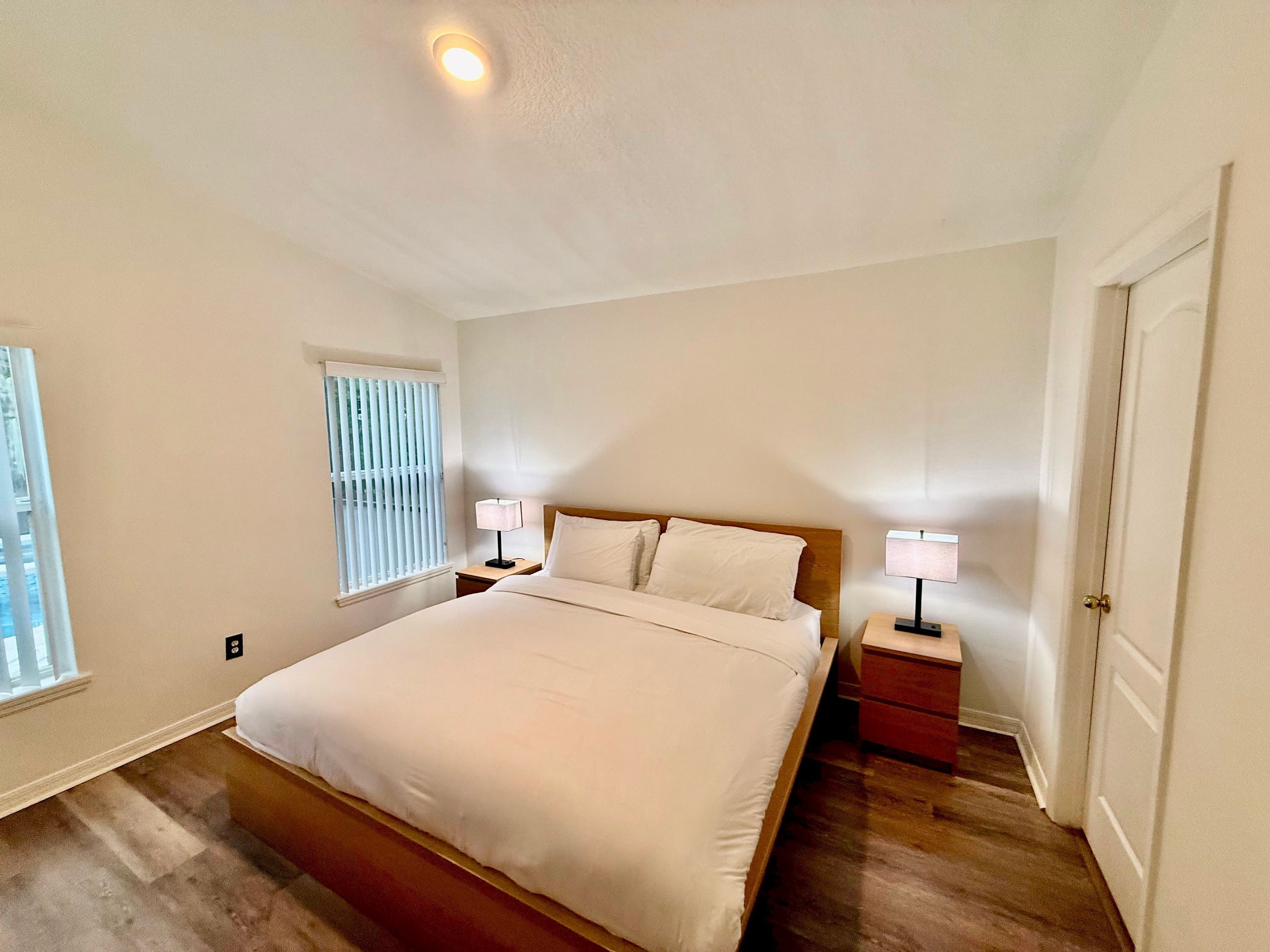 A bedroom with a bed, two bedside tables, a window with blinds, and a door.