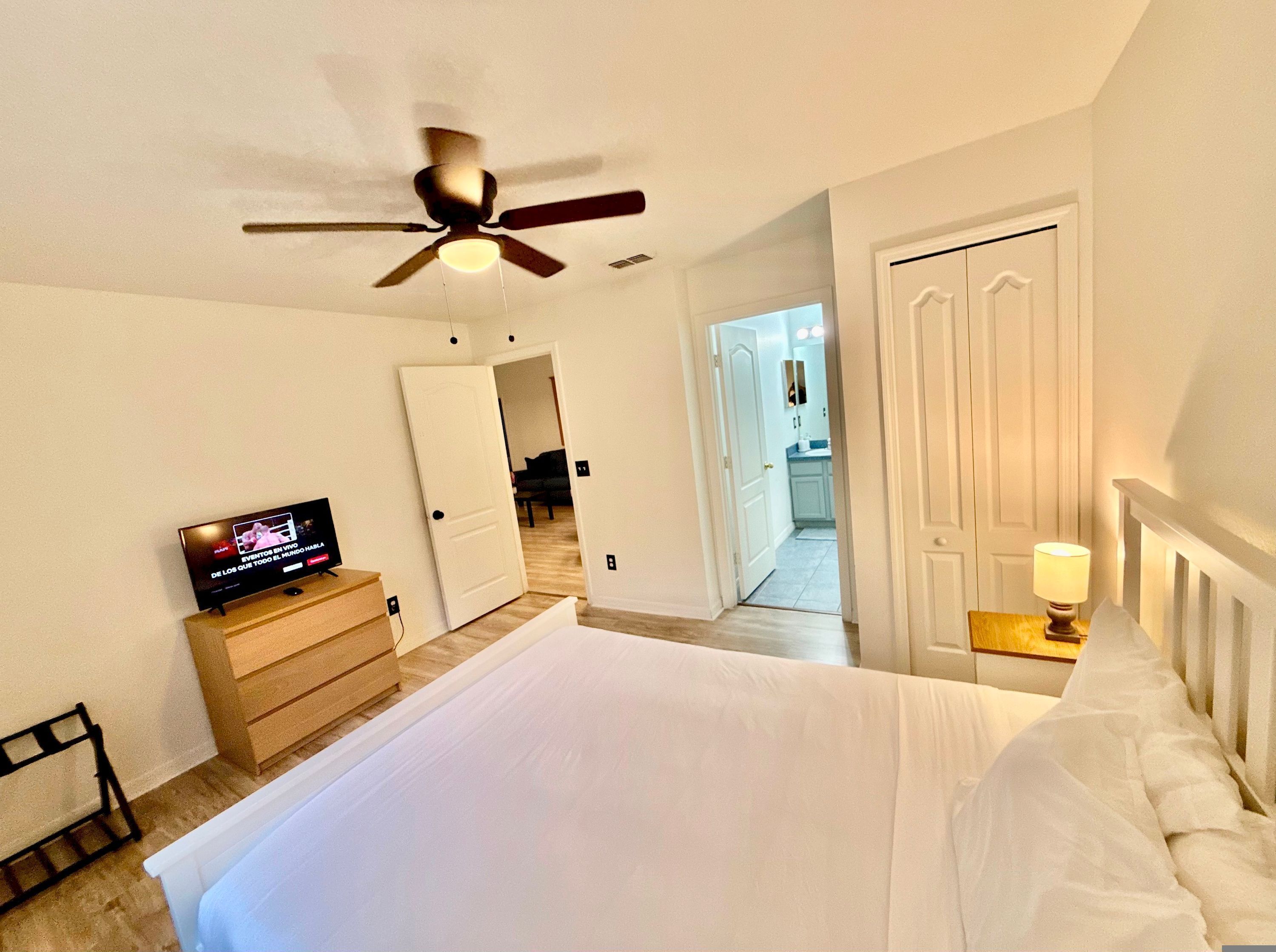 A bedroom with a bed, a ceiling fan, a TV, a wooden dresser, and a door leading to a bathroom.