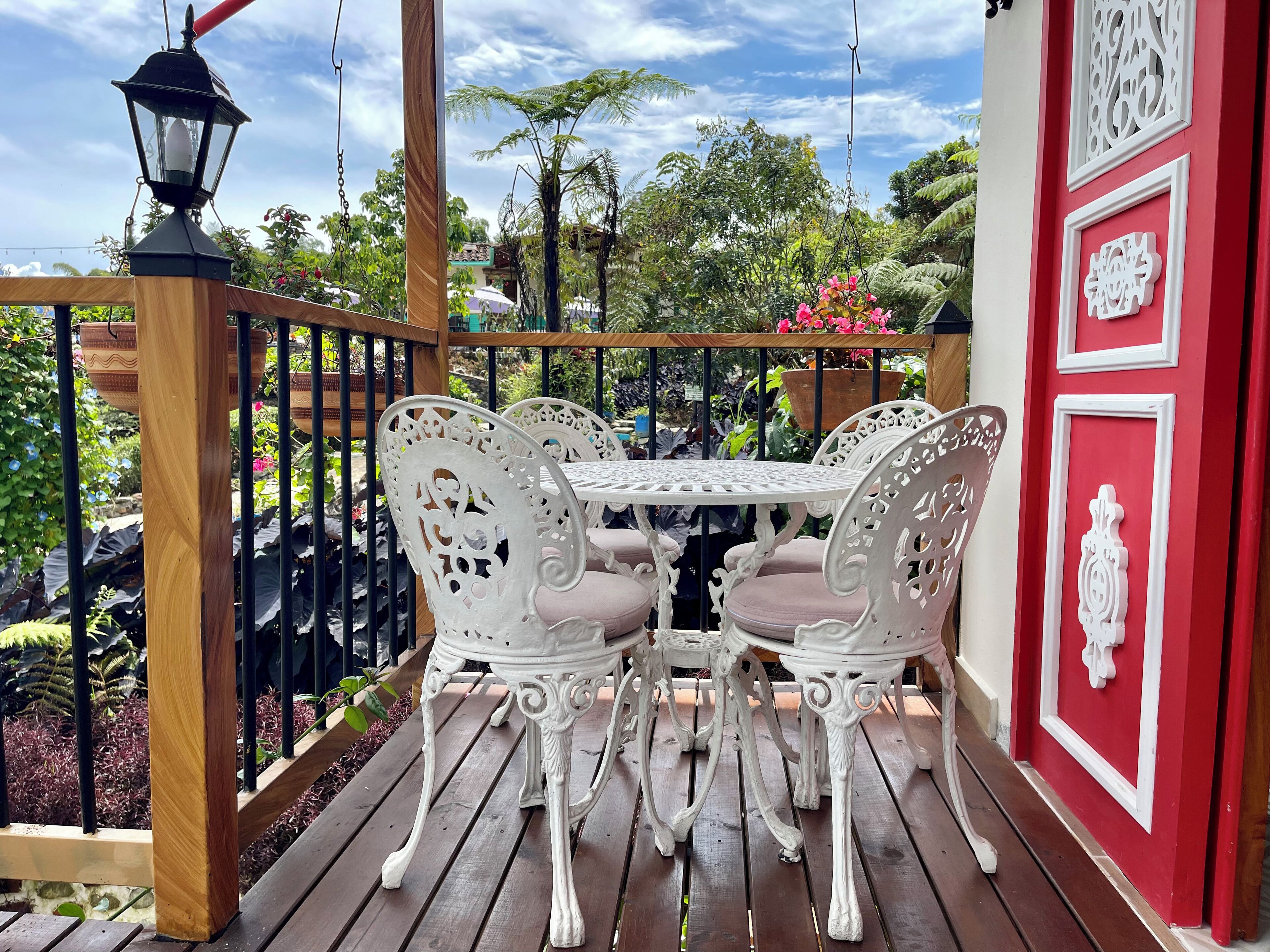 Een houten terras met witte, sierlijke stoelen en een tafel, omgeven door een zwarte metalen balustrade.