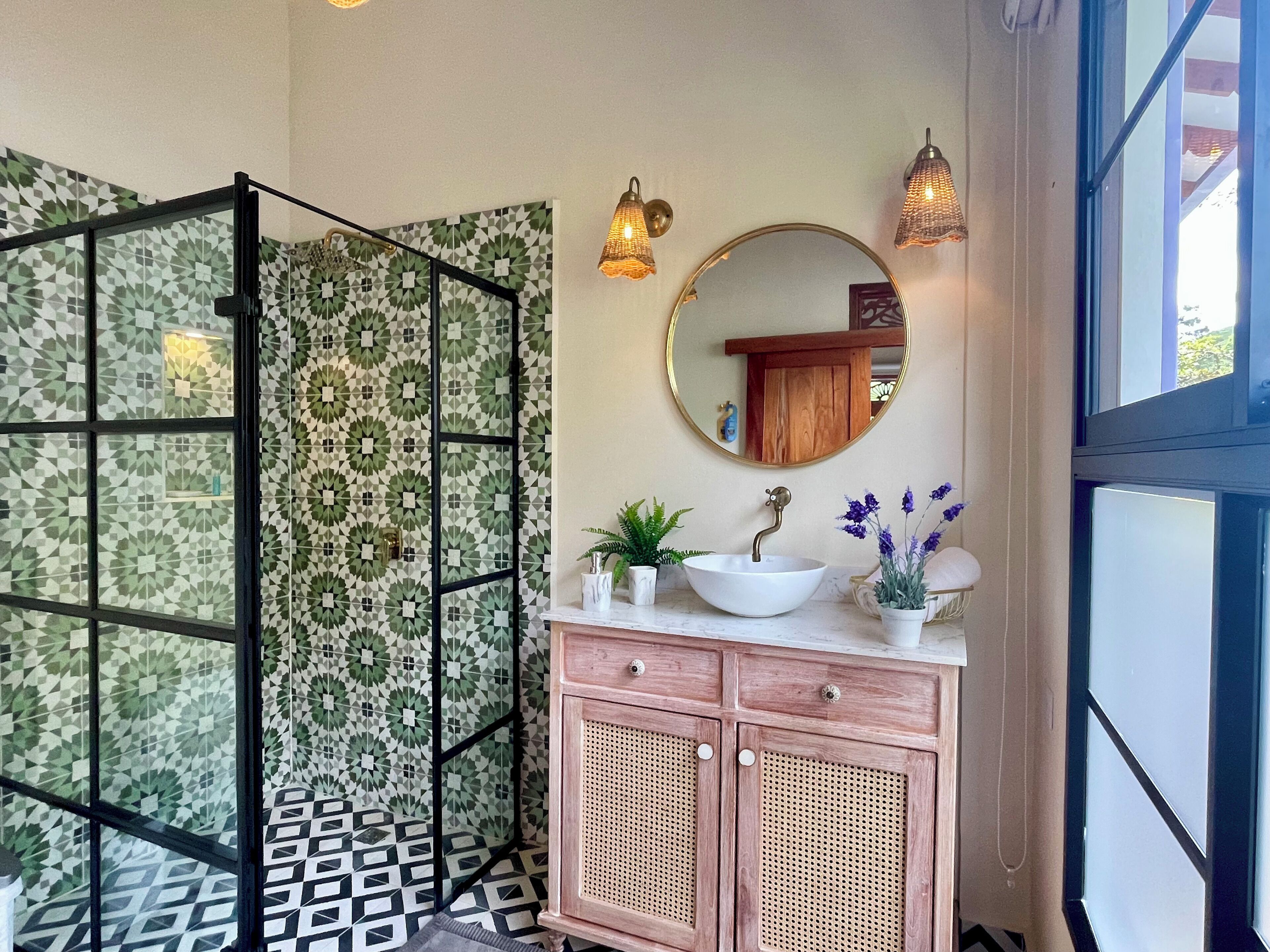 Un baño con pared de diseño geométrico, un espejo redondo, un lavabo blanco, un mueble con detalles de ratán y una zona de ducha con separador de metal negro.