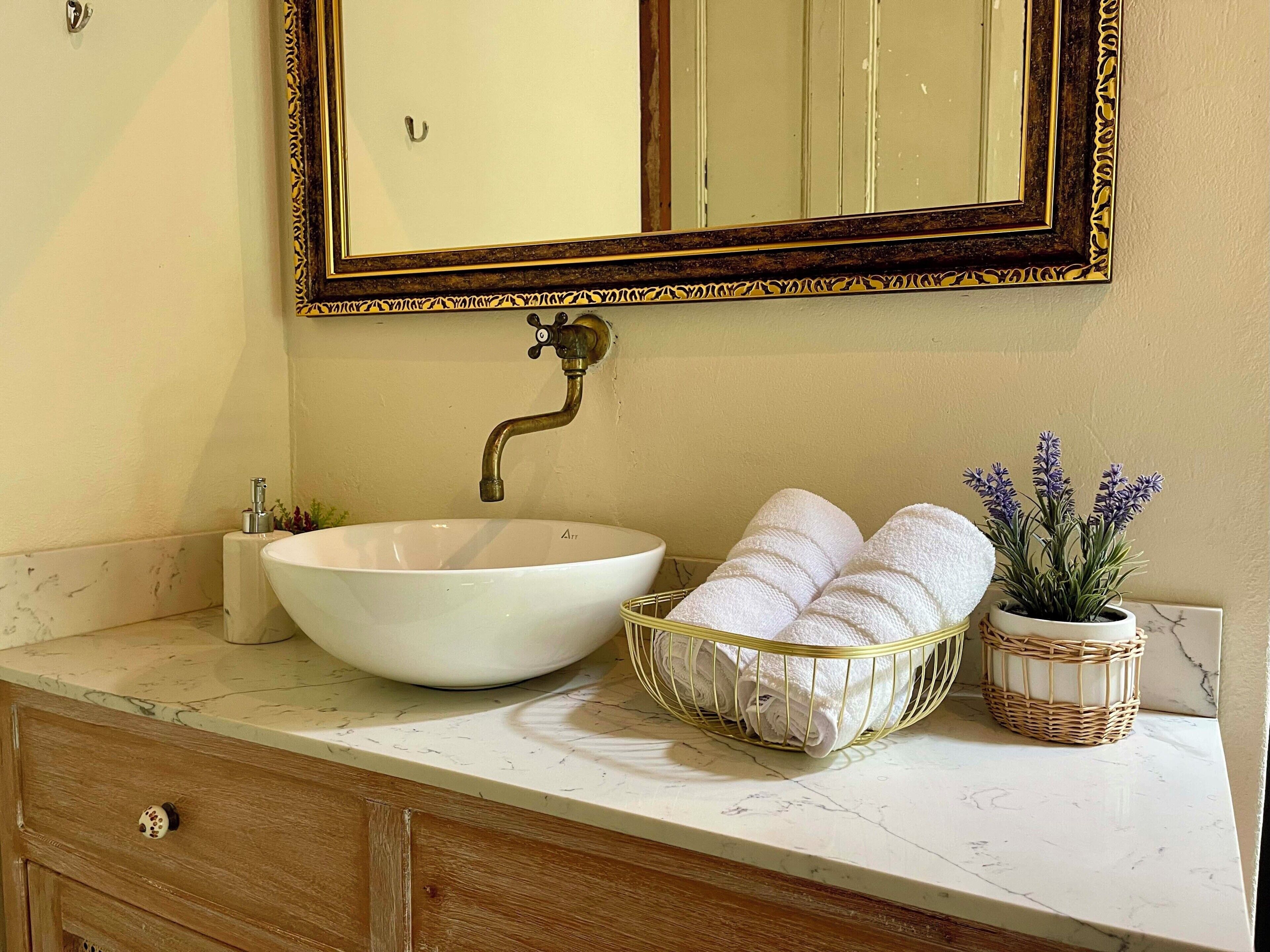 Un baño con un lavabo blanco, un espejo con marco dorado, un cesto para toallas y una planta de lavanda en maceta.
