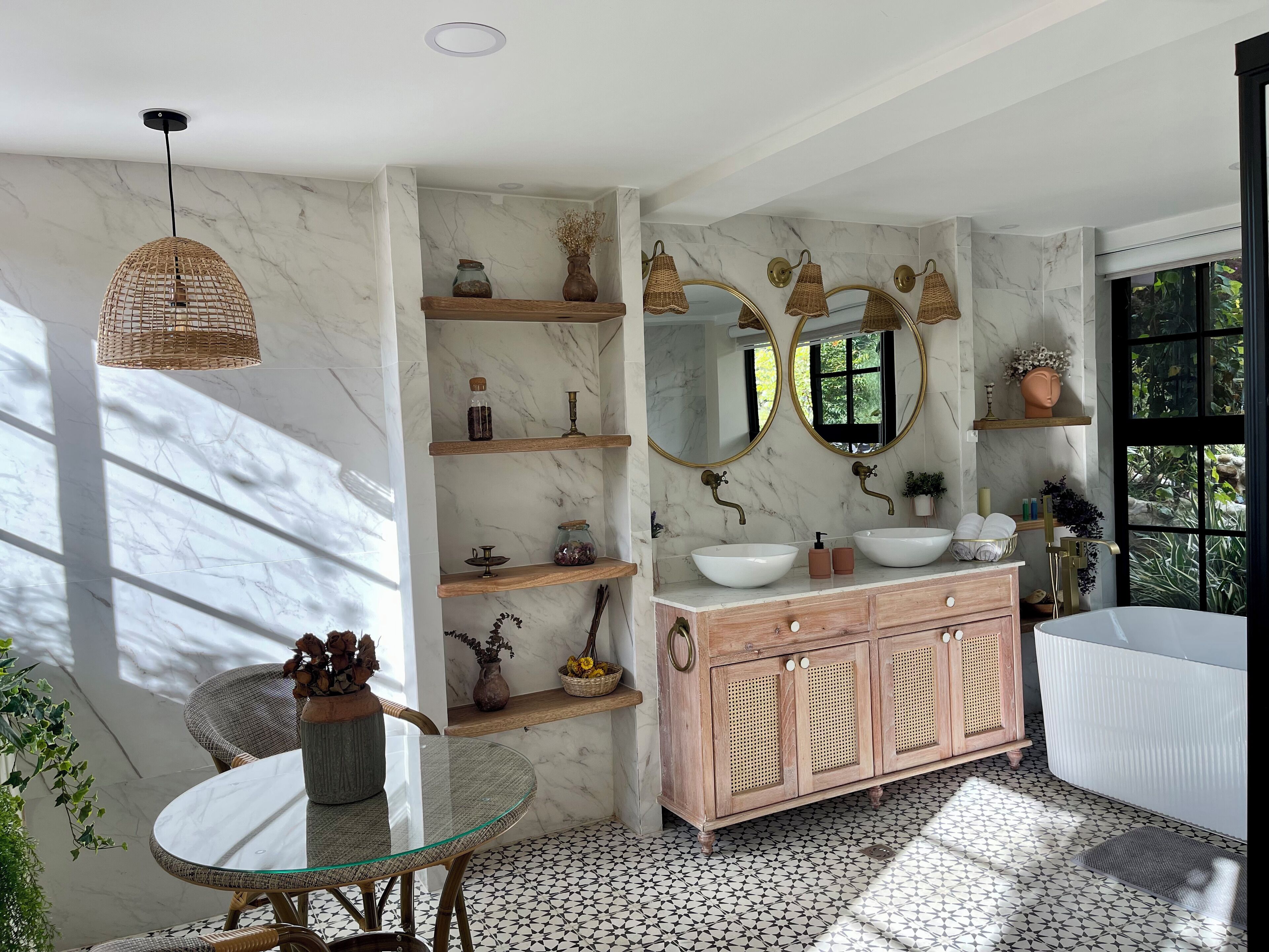 Un baño con pared de mármol, tocador de madera, mesita redonda de vidrio, lámparas colgantes de mimbre y un ventanal grande.