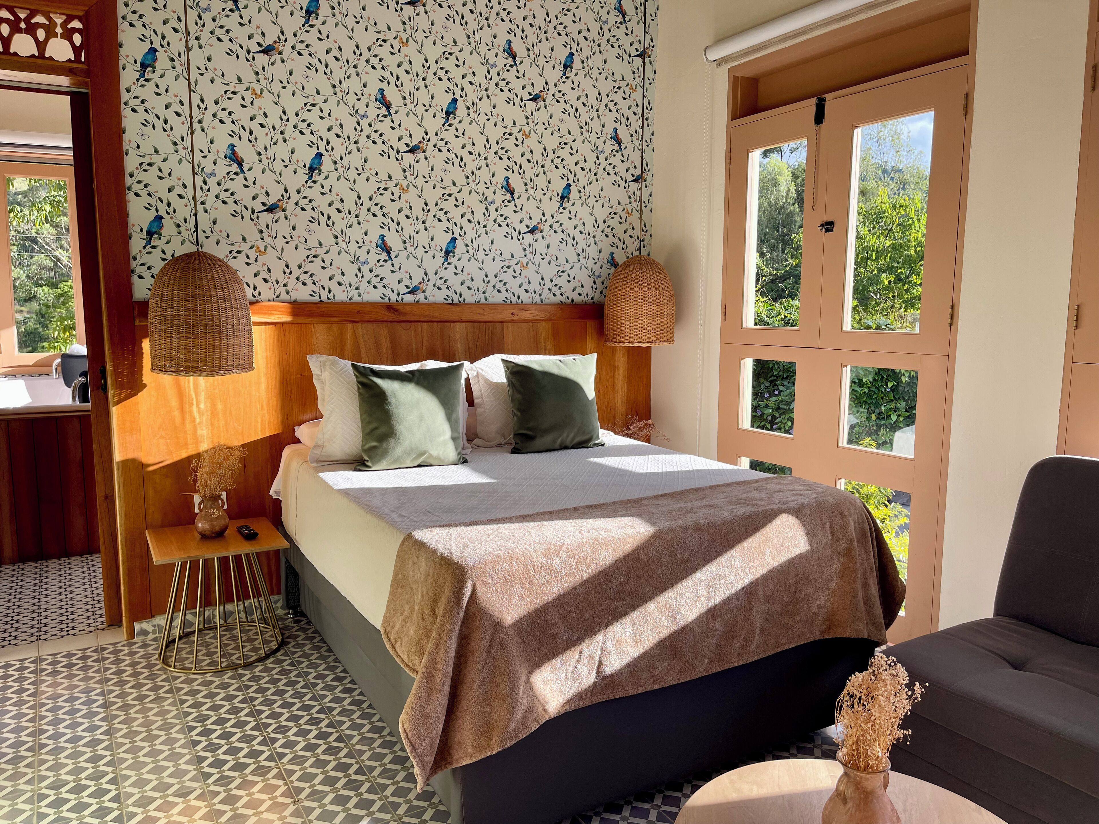 A bedroom with a wooden headboard, a bed with white bedding and green pillows, a bedside table with a vase and remote control, a patterned wall, and a window with a view of greenery.