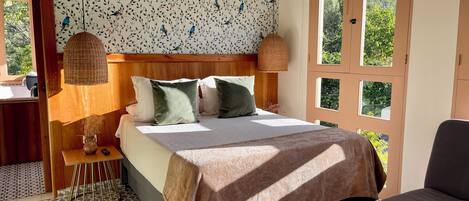 A bedroom with a wooden headboard, a bed with white bedding and green pillows, a bedside table with a vase and remote control, a patterned wall, and a window with a view of greenery.