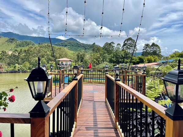Un puente de madera con barandillas y luces colgantes, que conduce a un paisaje verde exuberante con colinas y una masa de agua.