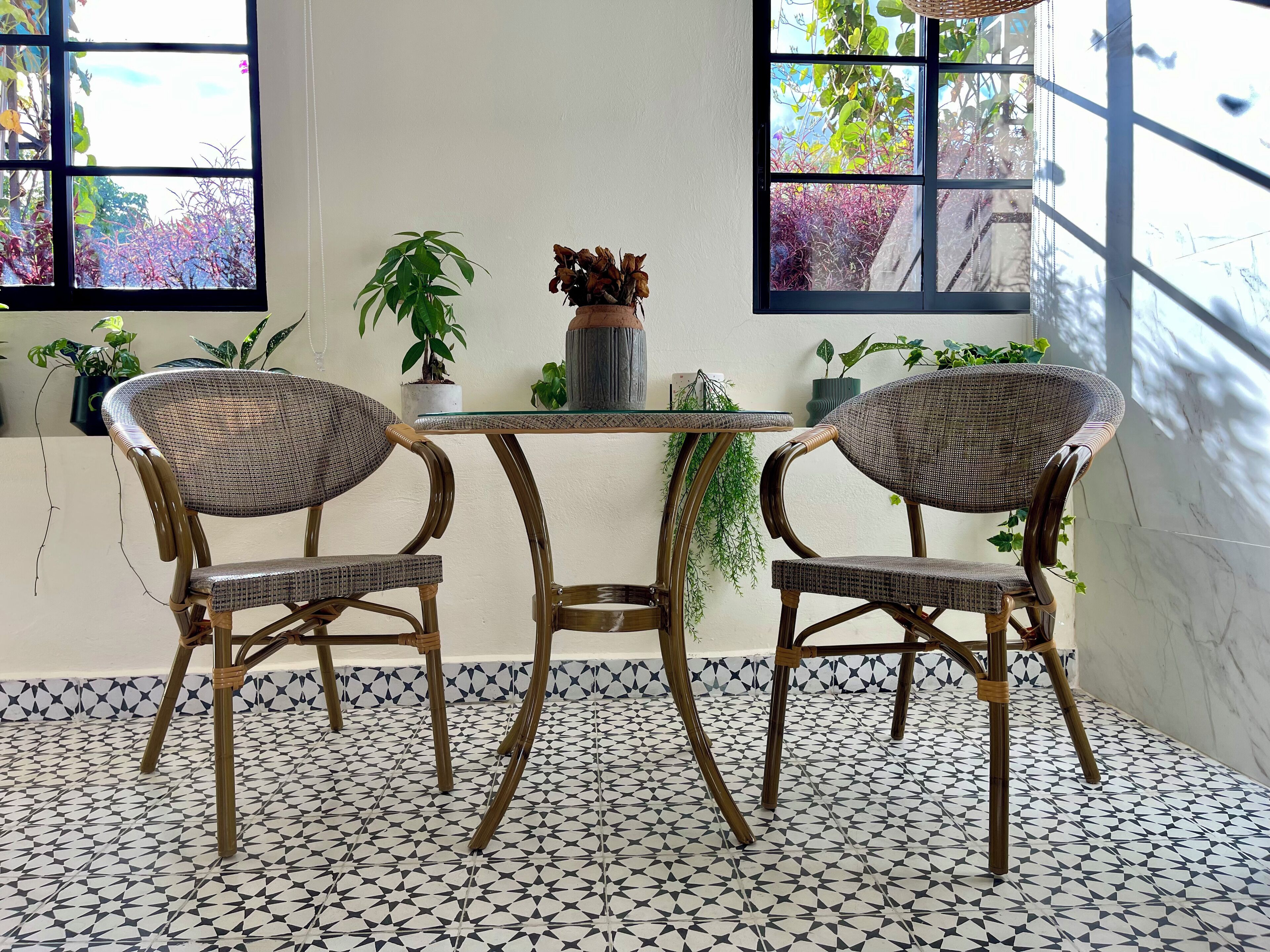 A room with a table and two wicker chairs, a patterned floor, and potted plants near the windows.