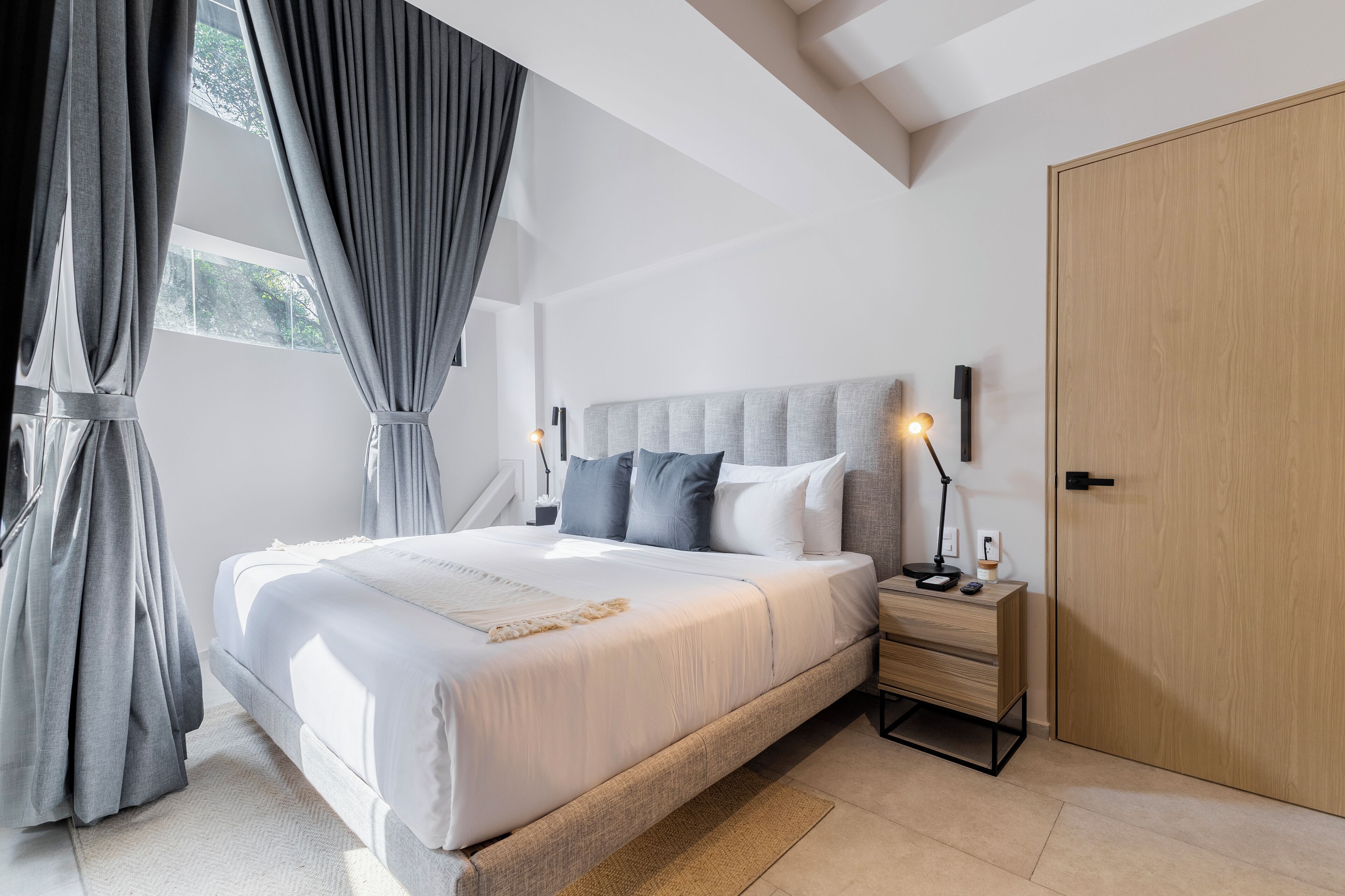 A modern bedroom with a large bed, grey curtains, a bedside table with a lamp, and a wooden wardrobe.