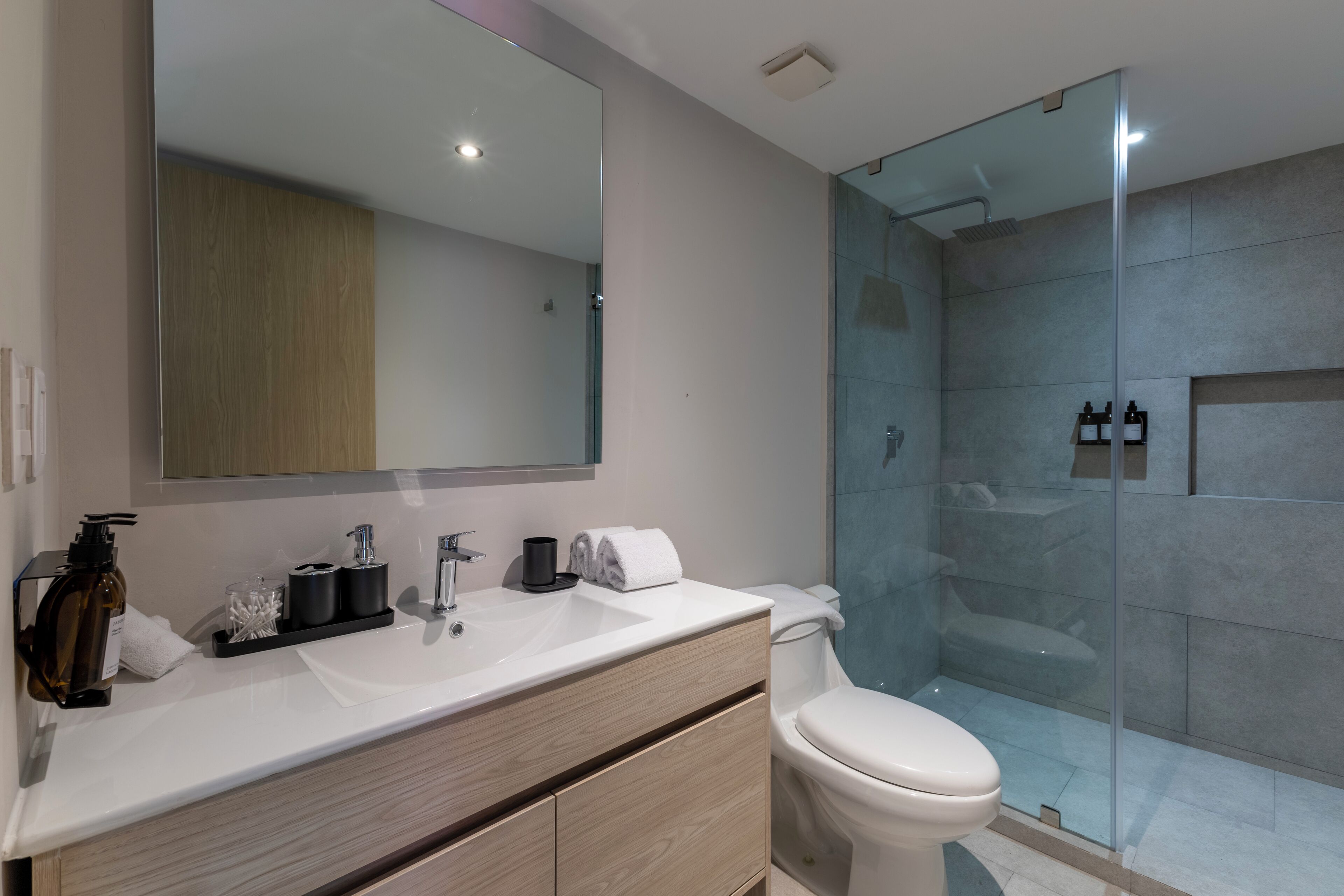 A modern bathroom with a large mirror, a white sink, a wooden vanity, a toilet, and a glass-enclosed shower area.