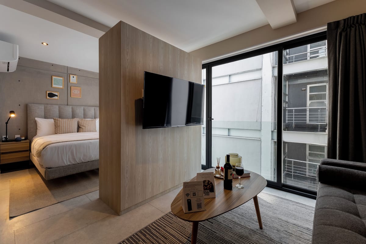 Una habitación de hotel moderna con una cama, un televisor de pantalla plana montado en la pared, una mesita de centro con una copa de vino y vistas a la ciudad a través de amplios ventanales.