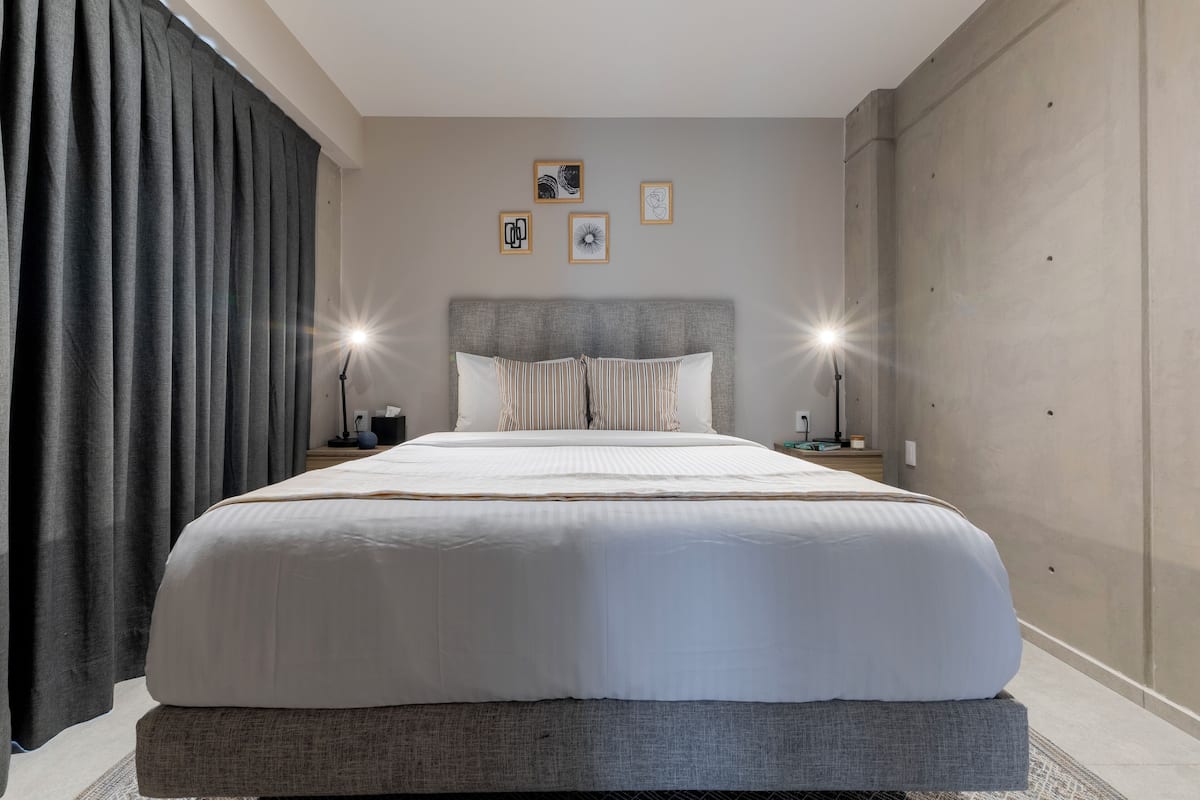 Una habitación de hotel moderna con una cama grande, dos mesitas de noche y un cabecero. Hay luces fijadas a la pared y tres cuadros enmarcados sobre la cama.