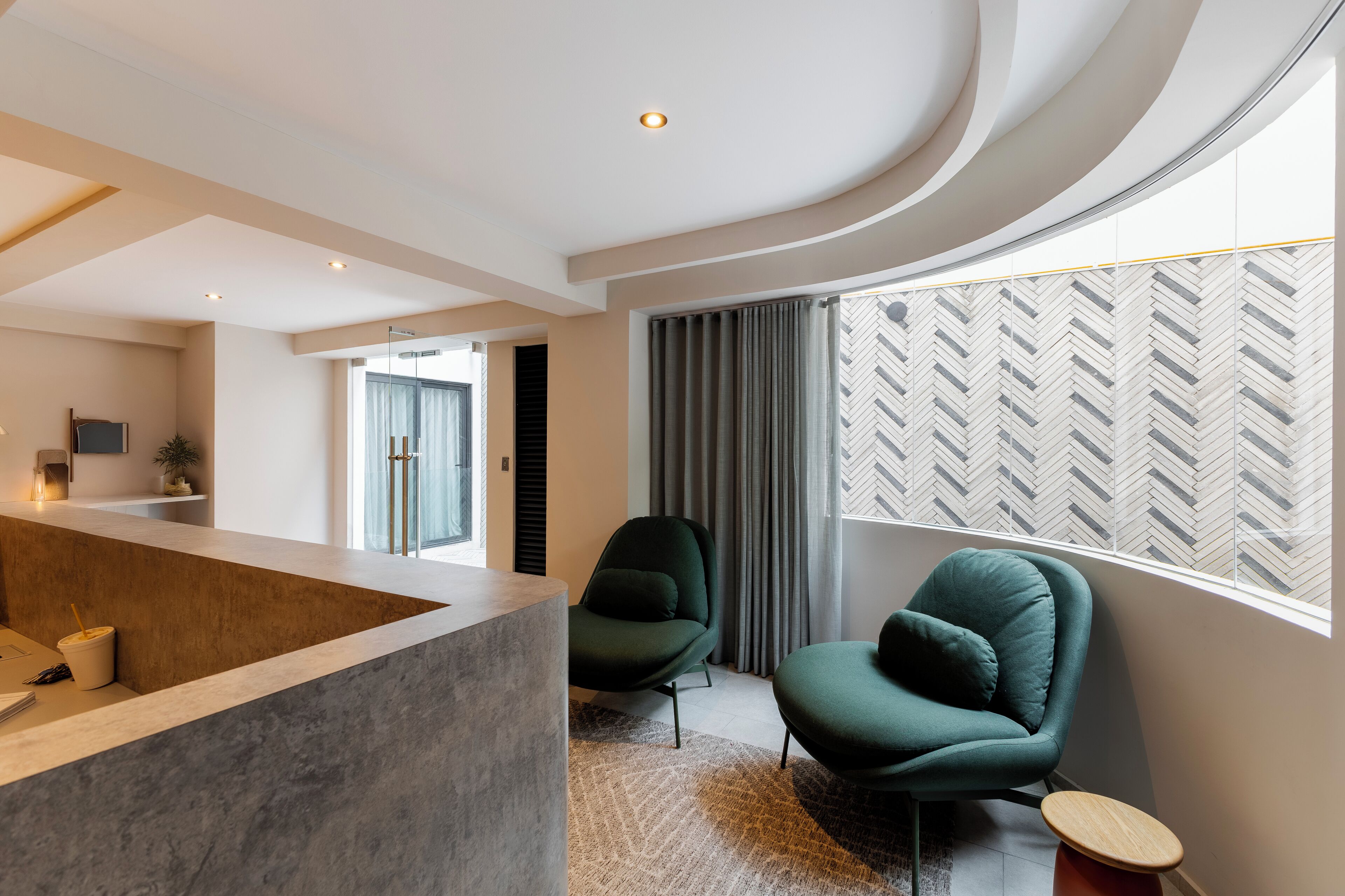 Un salon moderne doté d’un plafond incurvé, d’un coin salon avec deux fauteuils verts, une table basse et une grande fenêtre avec des stores à motifs.