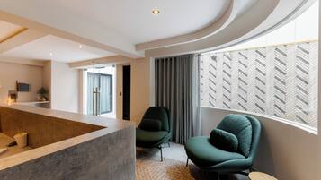 Un salon moderne doté d’un plafond incurvé, d’un coin salon avec deux fauteuils verts, une table basse et une grande fenêtre avec des stores à motifs.
