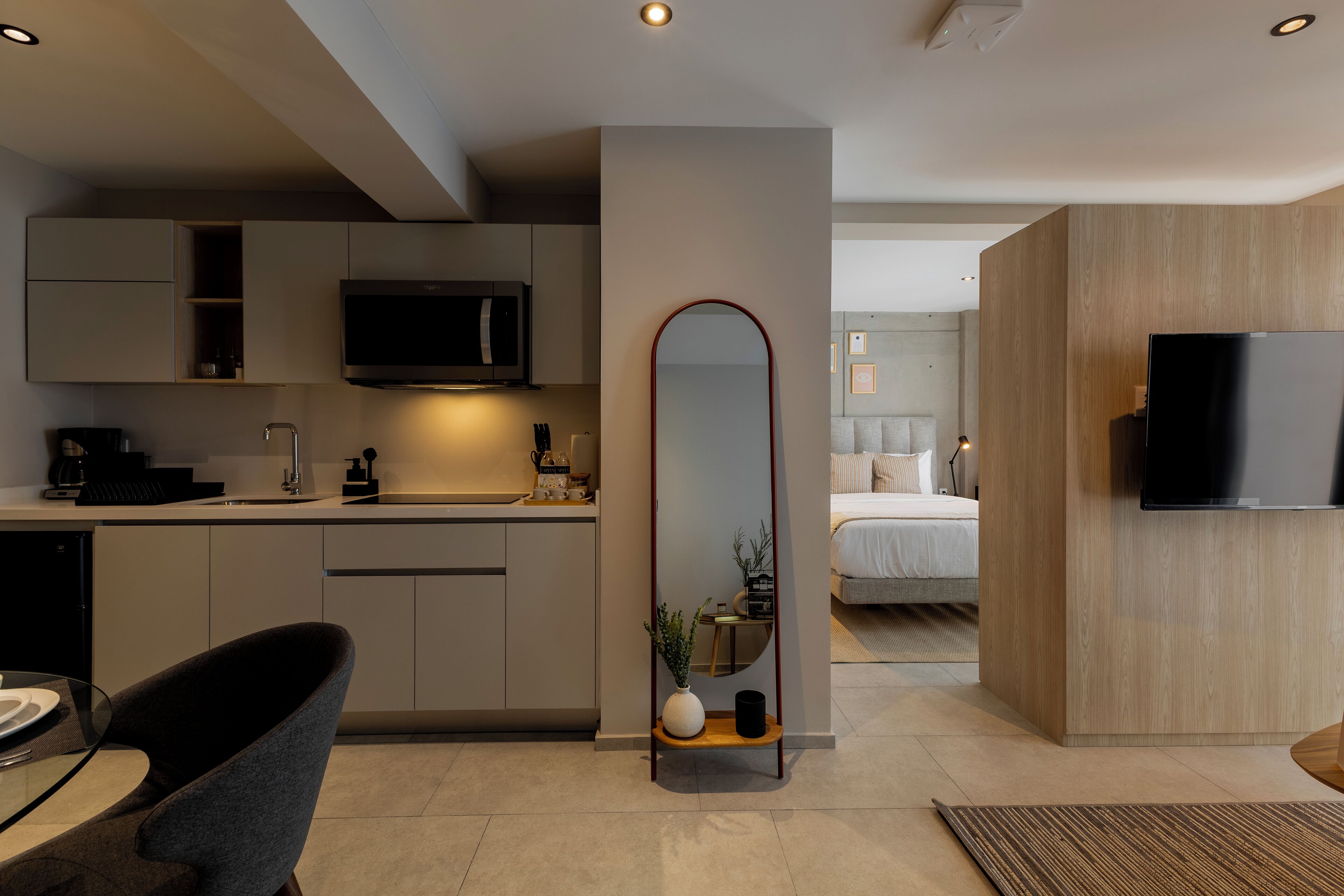 A modern kitchen with a built-in microwave, a dining area with a glass table and chairs, a large mirror, and a TV mounted on the wall.