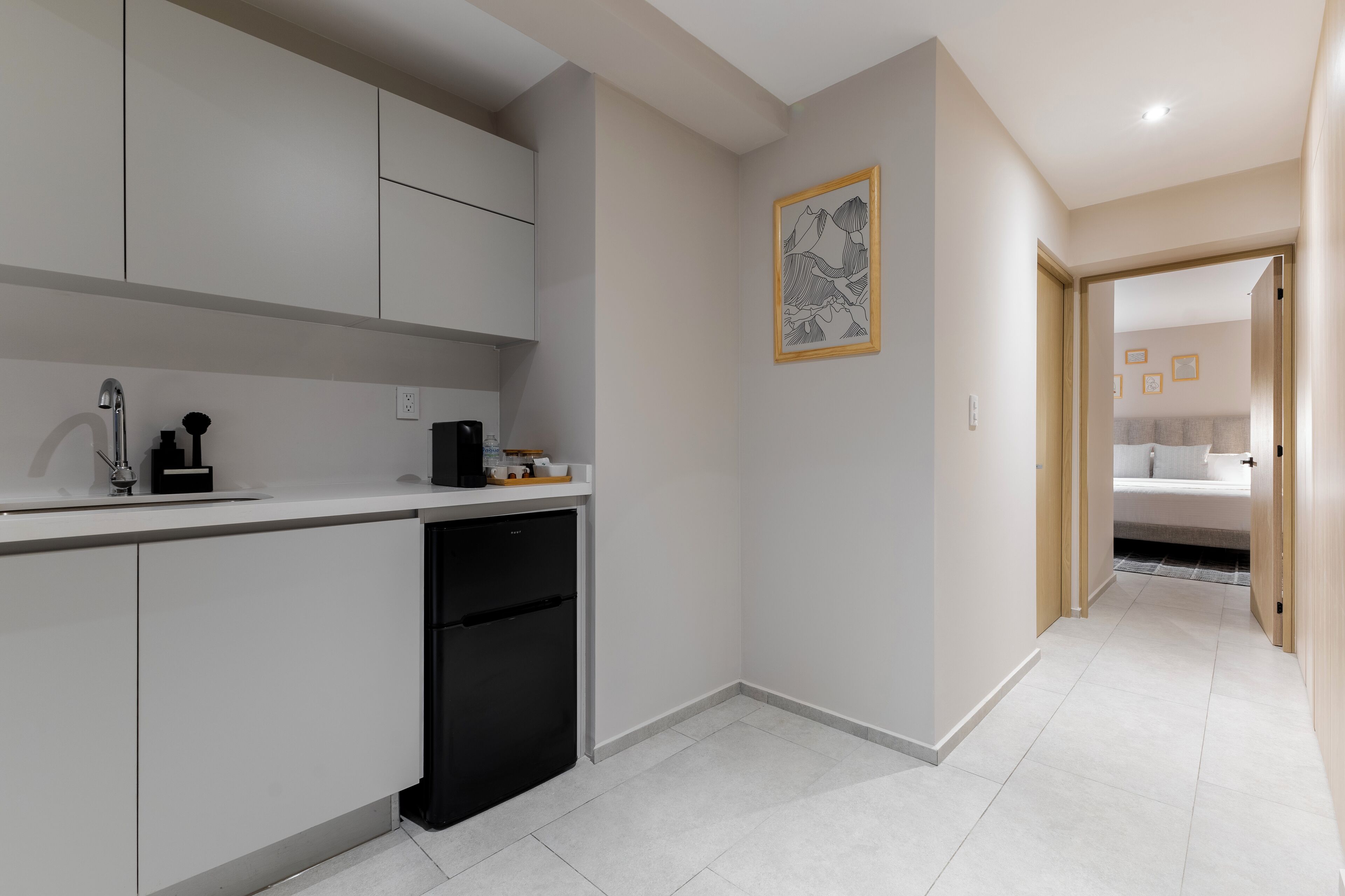 A modern kitchen with white cabinets, a black dishwasher, and a countertop with a coffee maker. A framed artwork is on the wall, and a hallway leads to a bedroom.