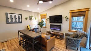 A modern living room with a wooden floor, a dining table with chairs, a sofa, a TV, and a ceiling fan. - Boho Getaway for Two, Stay near the New River Gorge and Glade Creek Farm (Cool Ridge)