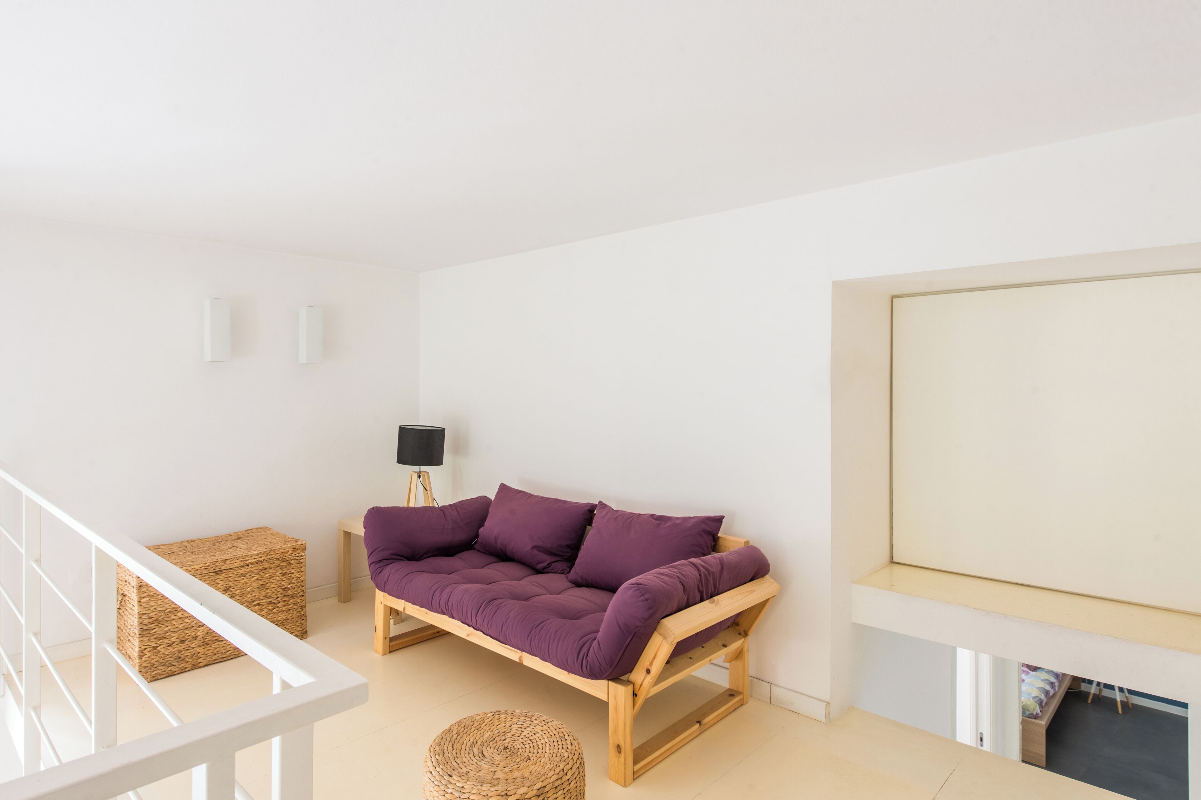 A minimalist living room with a wooden sofa, a small side table, a woven ottoman, and a black floor lamp.