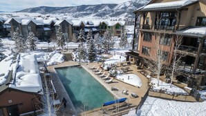 A resort with a pool surrounded by lounge chairs, snow-covered trees, and buildings with balconies.