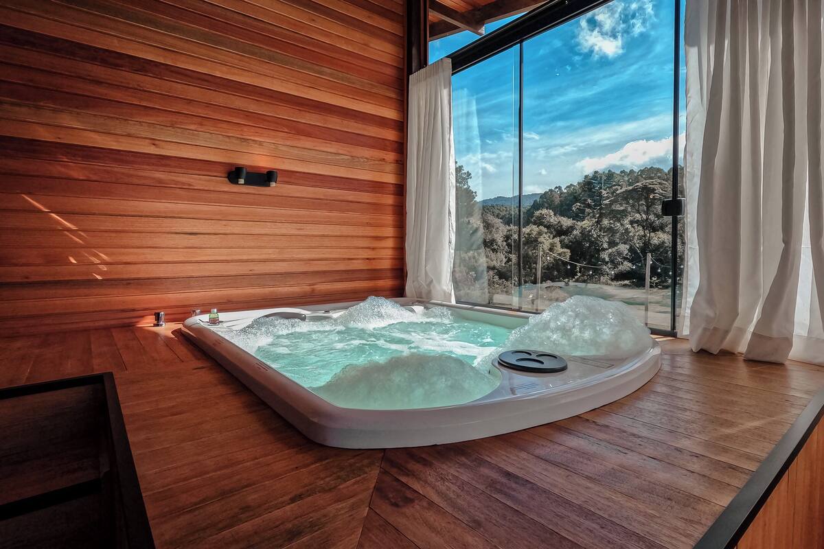 A hot tub with ice, wooden walls, and a view of trees and mountains through large windows.