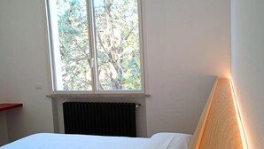Un dormitorio con cama, ventana con vistas a árboles, radiador y mesita de noche.