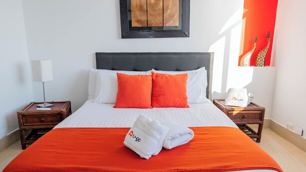 Ein ordentlich bezogenes Bett mit weißer und orangefarbener Bettwäsche, einem schwarzen Kopfteil und einem Paar weiße Handtücher mit einem Logo.