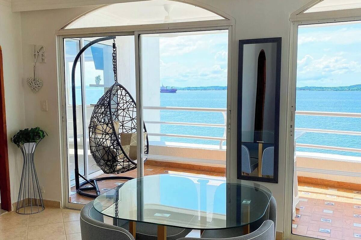 A dining area with a glass table and chairs, a hanging chair, and a view of the sea through large windows.