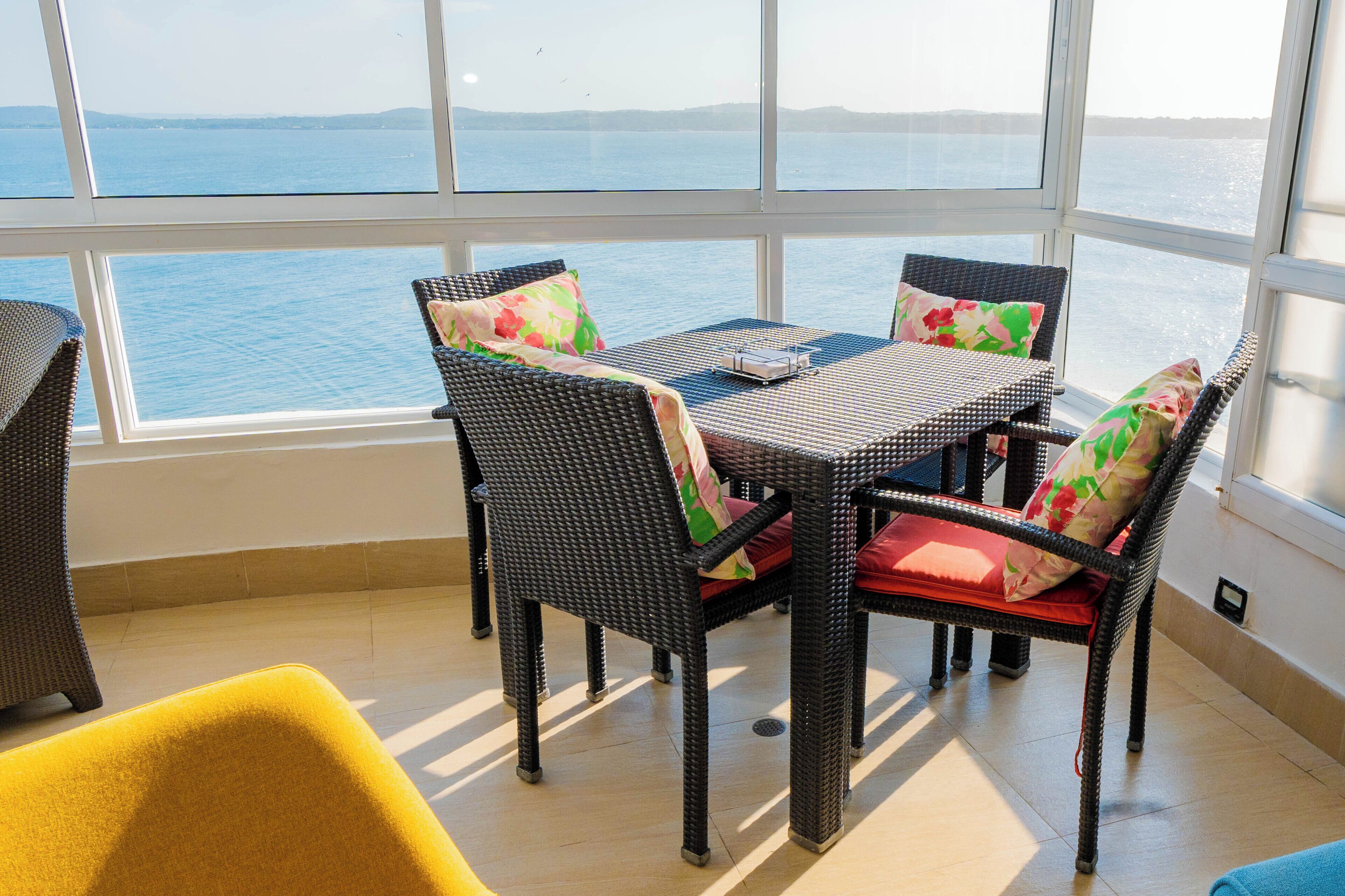 A room with a large window overlooking a body of water, furnished with wicker chairs and a table.