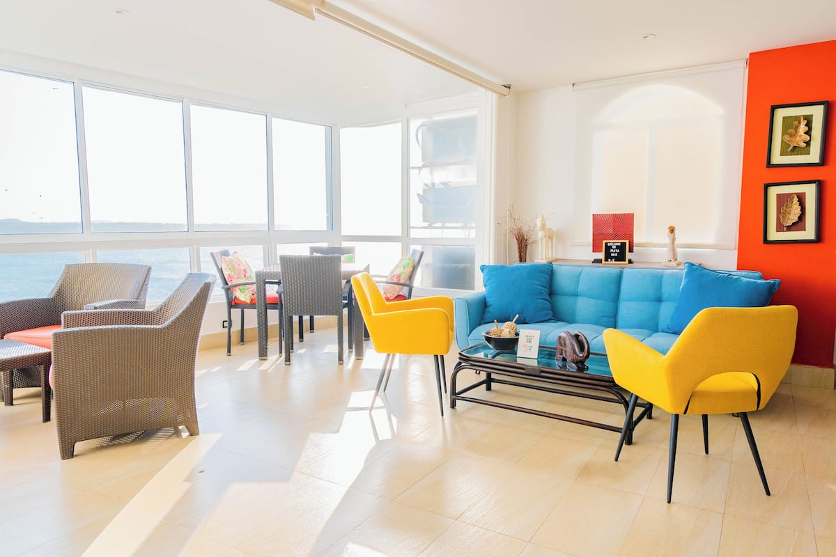 A bright living room with a blue sofa, yellow chairs, wicker furniture, and a view of the sea.