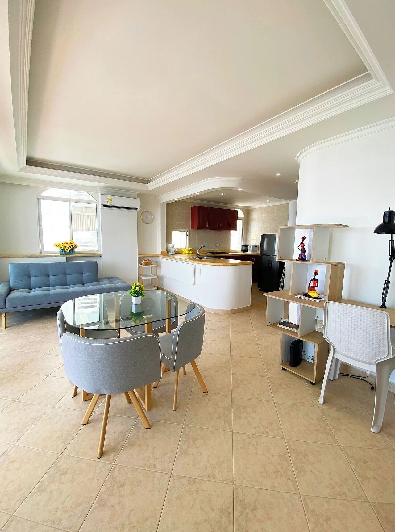 A modern living room with a glass dining table, grey chairs, a blue sofa, and a kitchen area with red cabinets and a white countertop.
