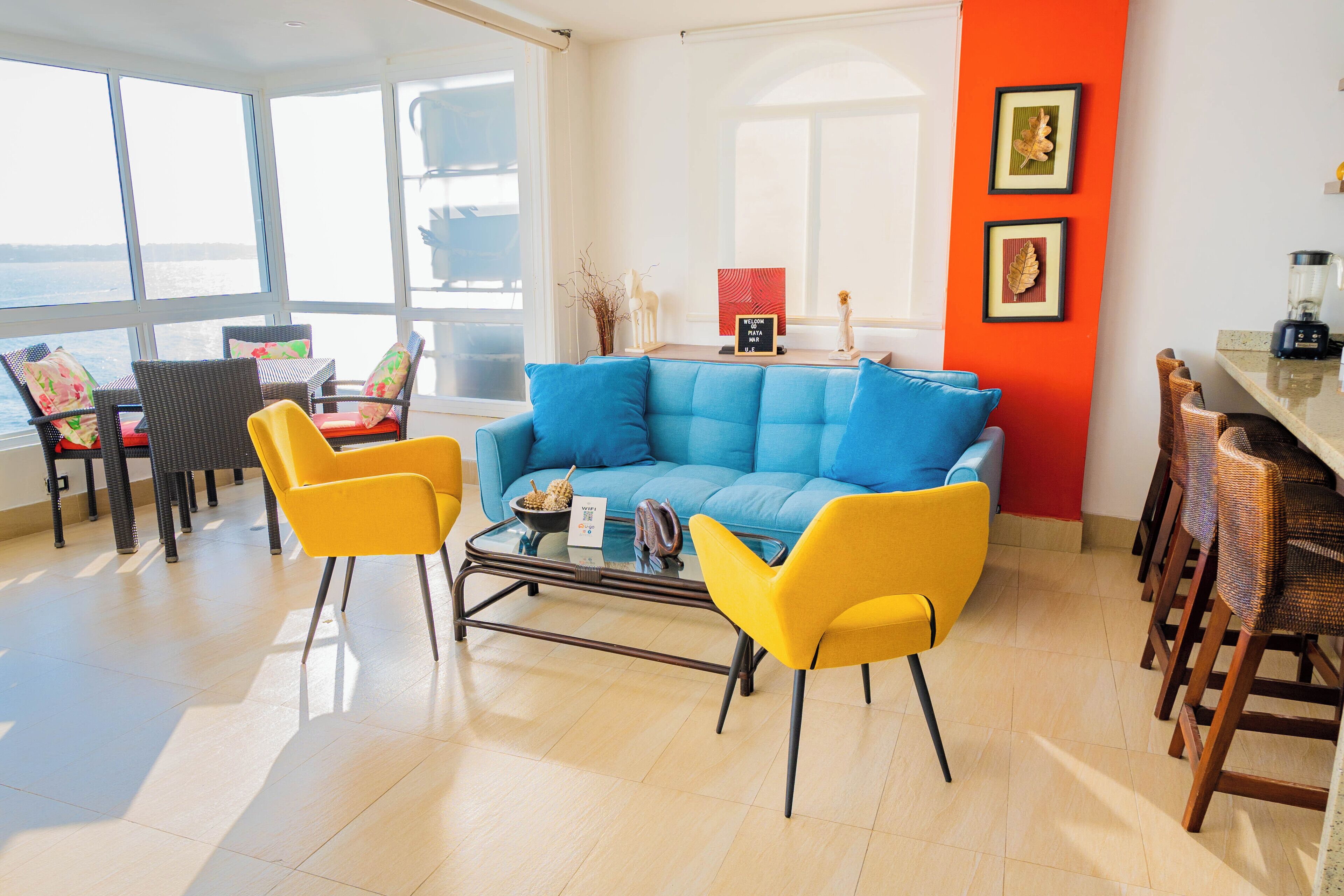 A bright living room with a blue sofa, yellow chairs, a glass coffee table, and a view of the sea through large windows.