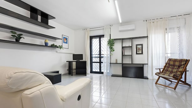 A modern living room with a sofa, a wooden rocking chair, and a TV. Shelves with plants and a picture frame are on the wall. - Casa Giovanna by Wonderful Italy (Bari)
