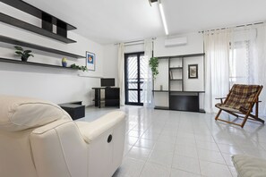 A modern living room with a sofa, a wooden rocking chair, and a TV. Shelves with plants and a picture frame are on the wall. - Casa Giovanna by Wonderful Italy (Bari)