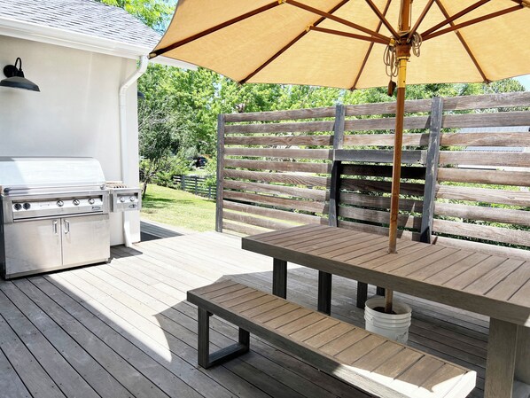 An outdoor patio with a grill, wooden table, bench, and umbrella.
