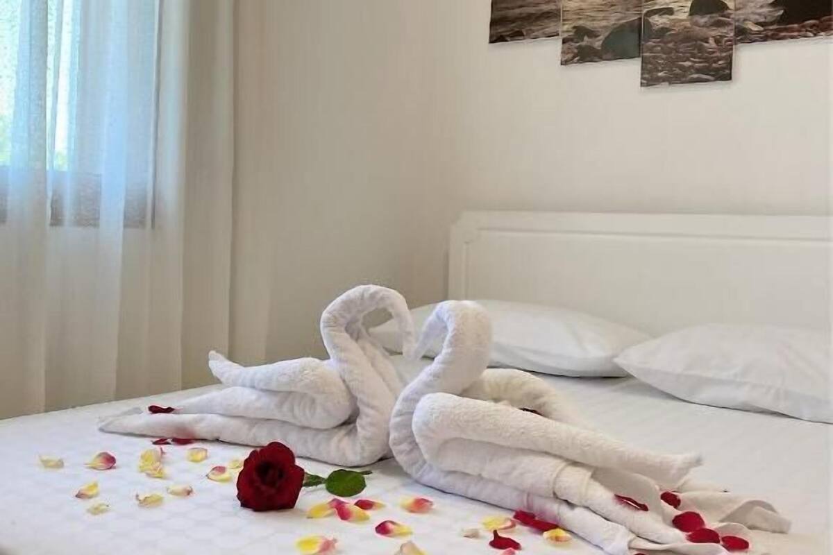 Cama bem arrumada com toalhas brancas dispostas em formato de cisne, uma rosa vermelha e pétalas de rosa espalhadas.