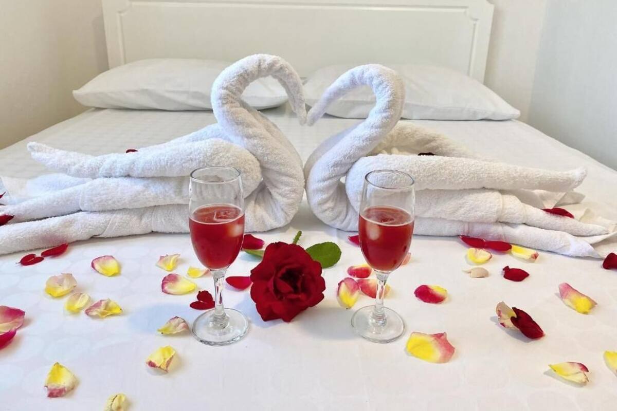 Uma cama com lençóis brancos, dois copos com bebida vermelha e uma rosa vermelha.