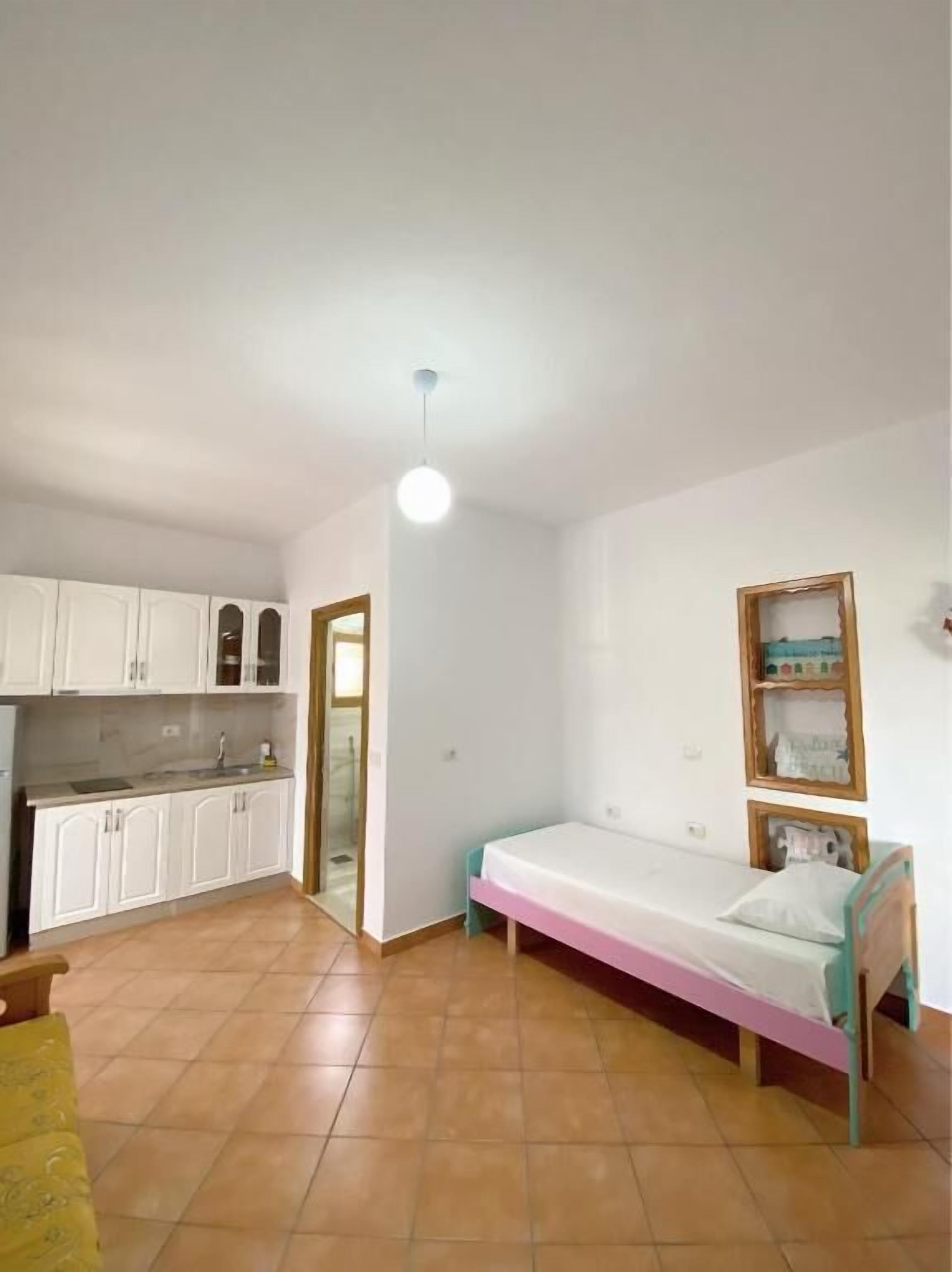 A small, bright room with a kitchenette, a single bed, and a door leading to another room.