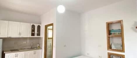 A small, bright room with a kitchenette, a single bed, and a door leading to another room.