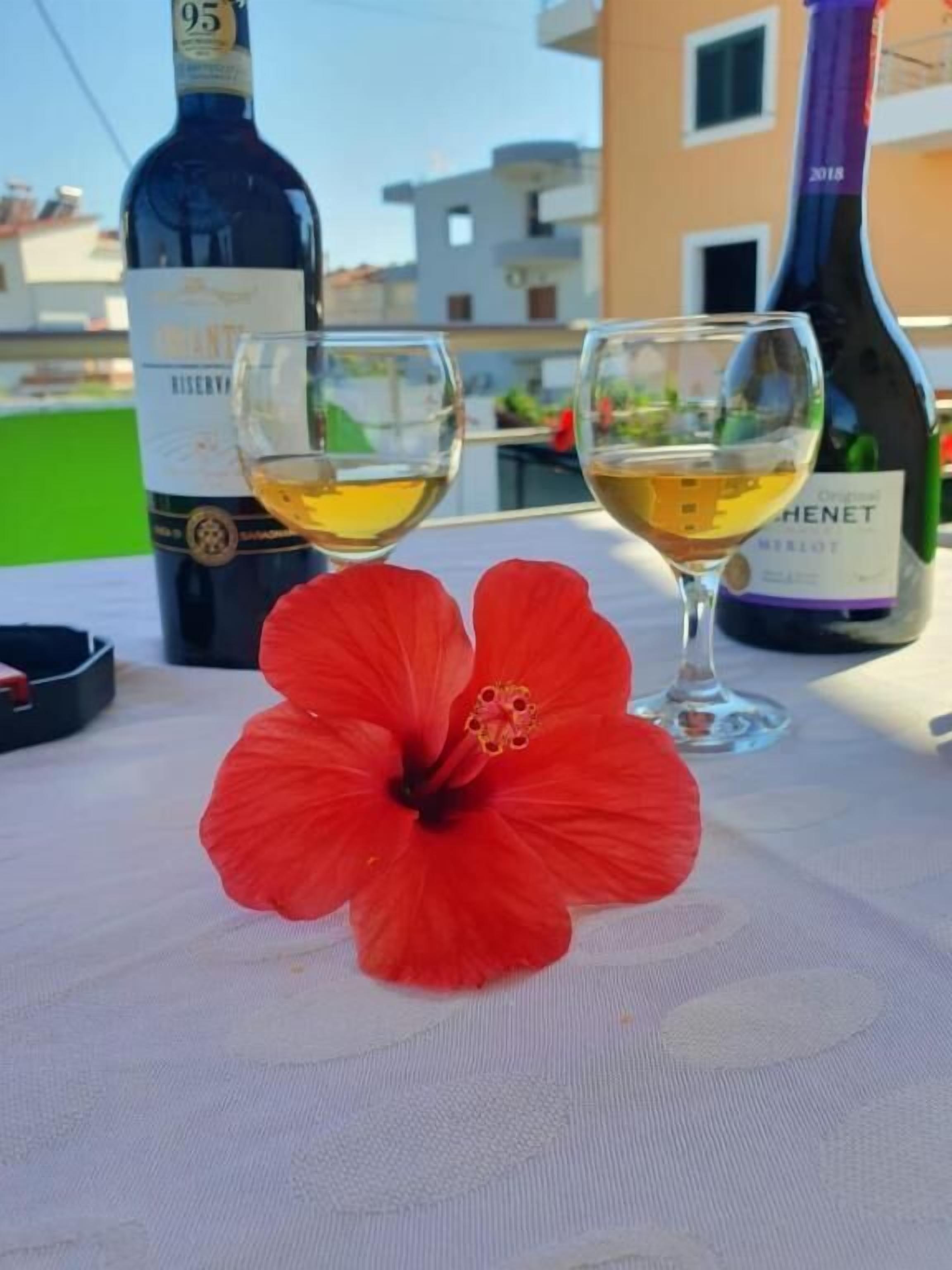 A table with two wine glasses filled with white wine, a bottle of wine, and a red hibiscus flower.