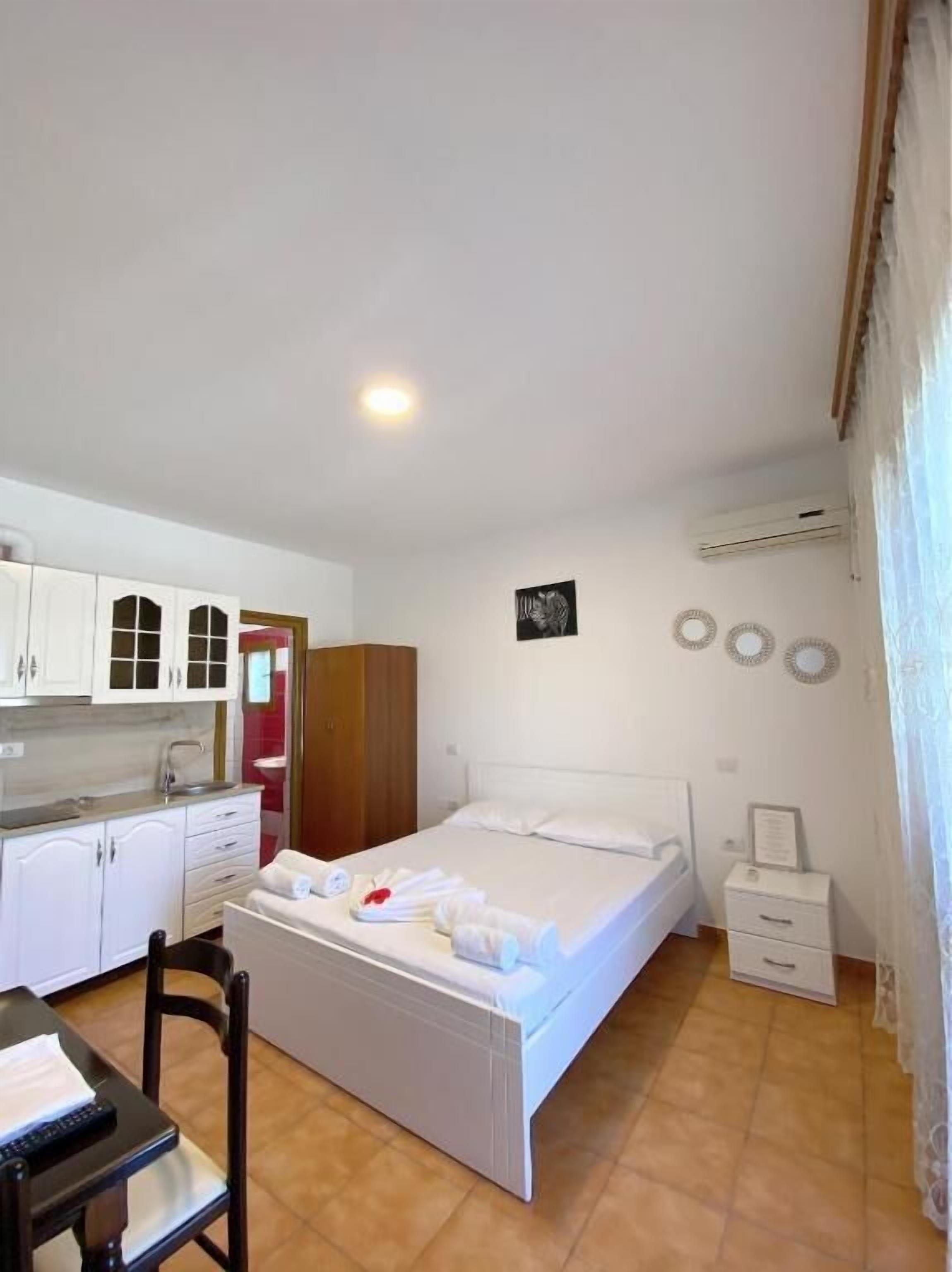A compact hotel room with a bed, a small desk, a chair, a kitchenette, and a bathroom.
