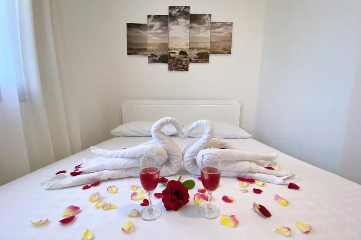 Cama com lençóis brancos, duas toalhas modeladas em forma de cisnes e dois copos com líquido vermelho.