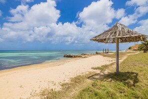 En strand med en stråtagsparasol, klart turkis vand og en sandstrand.