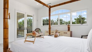 Uma cama com dossel e estrutura de madeira, uma pequena mesa de madeira com uma planta e um abajur, uma cômoda com gavetas e uma janela grande com vista para árvores.