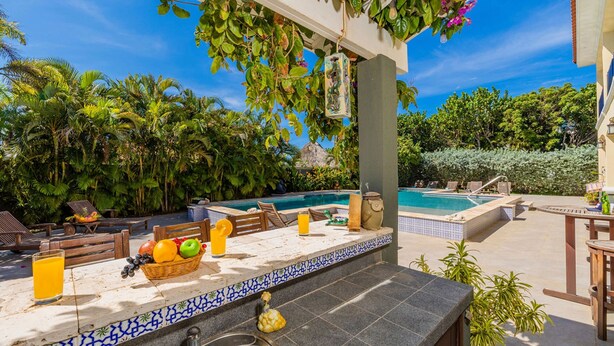 Uma área na piscina com um bar revestido em azulejos, espreguiçadeiras e vista para a piscina e vegetação tropical.