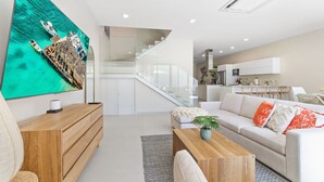 Ein modernes Wohnzimmer mit einem großen Flachbildfernseher, einem Holztisch, einem weißen Sofa mit orangefarbenen und weißen Kissen und einer Treppe, die zu einer oberen Etage führt.